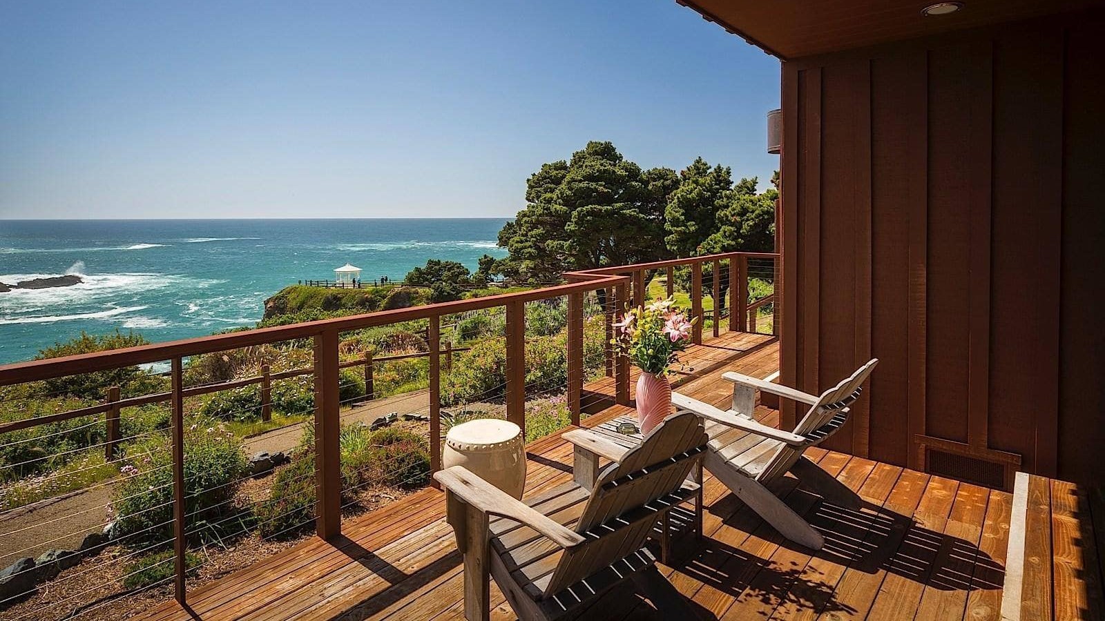 A vibrant dog park bathed in sunlight features abundant greenery with mature trees, offering ample shade for leisurely outdoor activities. In the distance is a well-equipped playground that presents an additional activity area. An inviting picnic table spreads out nearby, ideal for enjoying meals al fresco or taking relaxing breaks from play. A neatly-placed trash bin aids in maintaining the park's cleanliness for everyone's enjoyment. Dominating the scene is a generously wide dirt path gracefully curving through the expanse of grass, promising exploratory walks and lively fetch games.