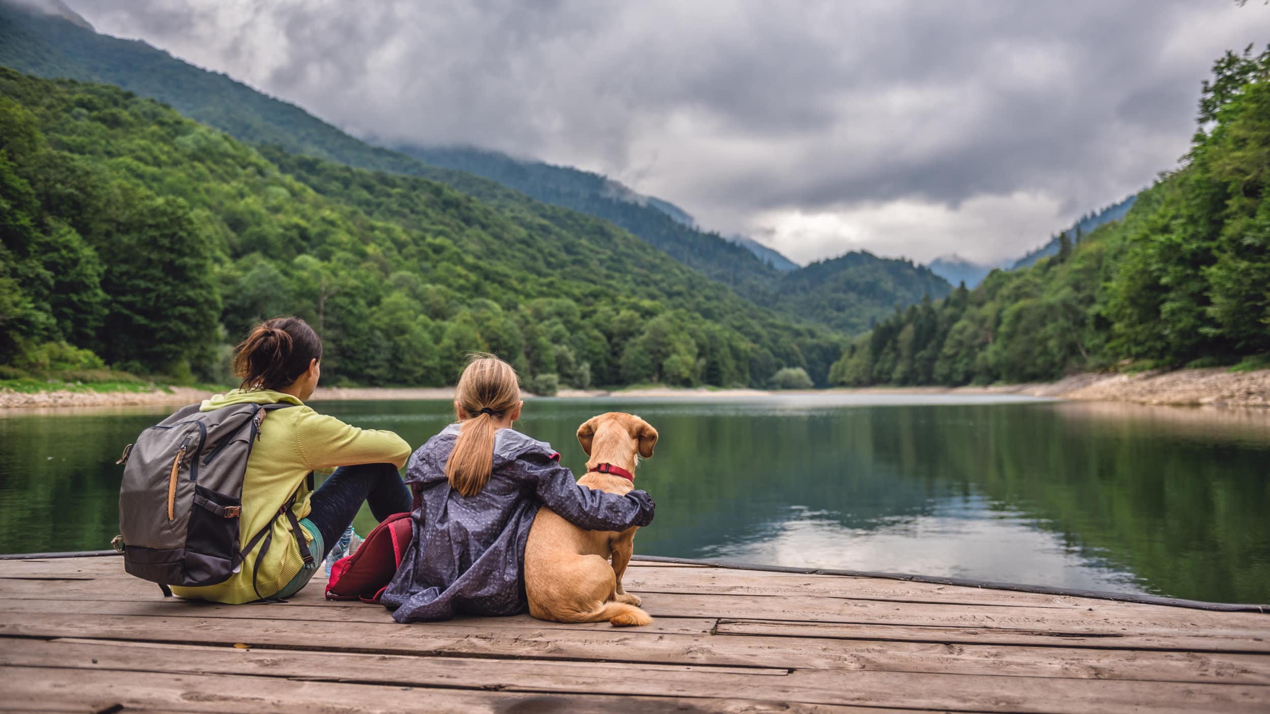 Mother and daughter with a dog resting on a pier looking at lake