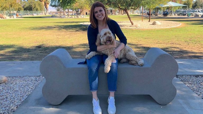 A cheerful woman, comfortably seated on a sturdy concrete bench amidst the vibrant green hues of an inviting park. Her hand firmly grips the leash, attached to an elated canine companion who is captivated by the sights and sounds around them. - Dogtrekker