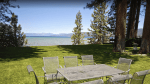 A vibrant dog park bathed in sunlight features abundant greenery with mature trees, offering ample shade for leisurely outdoor activities. In the distance is a well-equipped playground that presents an additional activity area. An inviting picnic table spreads out nearby, ideal for enjoying meals al fresco or taking relaxing breaks from play. A neatly-placed trash bin aids in maintaining the park's cleanliness for everyone's enjoyment. Dominating the scene is a generously wide dirt path gracefully curving through the expanse of grass, promising exploratory walks and lively fetch games.