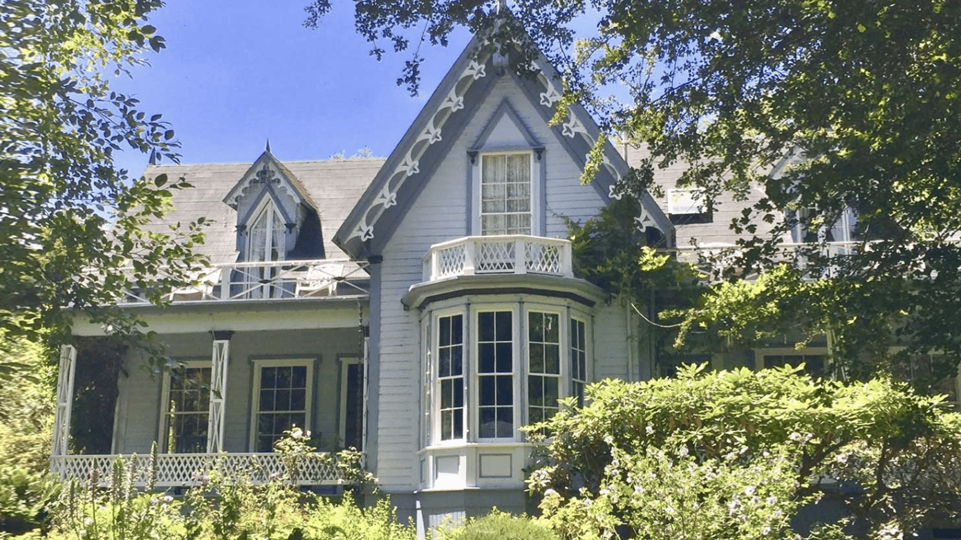 A vibrant dog park bathed in sunlight features abundant greenery with mature trees, offering ample shade for leisurely outdoor activities. In the distance is a well-equipped playground that presents an additional activity area. An inviting picnic table spreads out nearby, ideal for enjoying meals al fresco or taking relaxing breaks from play. A neatly-placed trash bin aids in maintaining the park's cleanliness for everyone's enjoyment. Dominating the scene is a generously wide dirt path gracefully curving through the expanse of grass, promising exploratory walks and lively fetch games.