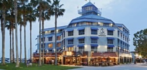 A vibrant dog park bathed in sunlight features abundant greenery with mature trees, offering ample shade for leisurely outdoor activities. In the distance is a well-equipped playground that presents an additional activity area. An inviting picnic table spreads out nearby, ideal for enjoying meals al fresco or taking relaxing breaks from play. A neatly-placed trash bin aids in maintaining the park's cleanliness for everyone's enjoyment. Dominating the scene is a generously wide dirt path gracefully curving through the expanse of grass, promising exploratory walks and lively fetch games.