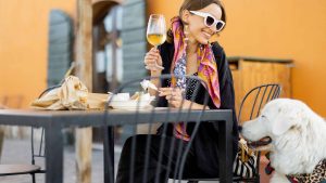 A vibrant dog park bathed in sunlight features abundant greenery with mature trees, offering ample shade for leisurely outdoor activities. In the distance is a well-equipped playground that presents an additional activity area. An inviting picnic table spreads out nearby, ideal for enjoying meals al fresco or taking relaxing breaks from play. A neatly-placed trash bin aids in maintaining the park's cleanliness for everyone's enjoyment. Dominating the scene is a generously wide dirt path gracefully curving through the expanse of grass, promising exploratory walks and lively fetch games.