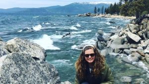 A vibrant dog park bathed in sunlight features abundant greenery with mature trees, offering ample shade for leisurely outdoor activities. In the distance is a well-equipped playground that presents an additional activity area. An inviting picnic table spreads out nearby, ideal for enjoying meals al fresco or taking relaxing breaks from play. A neatly-placed trash bin aids in maintaining the park's cleanliness for everyone's enjoyment. Dominating the scene is a generously wide dirt path gracefully curving through the expanse of grass, promising exploratory walks and lively fetch games.