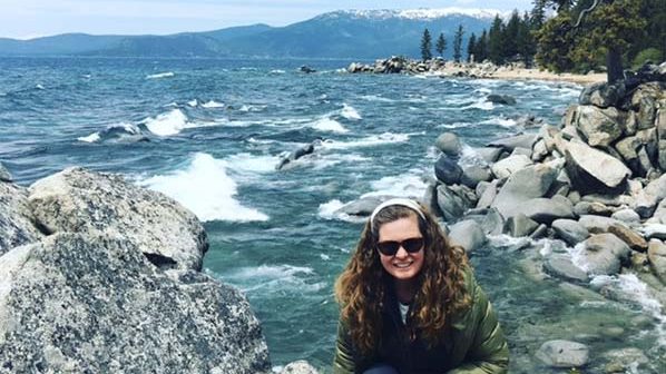 A vibrant dog park bathed in sunlight features abundant greenery with mature trees, offering ample shade for leisurely outdoor activities. In the distance is a well-equipped playground that presents an additional activity area. An inviting picnic table spreads out nearby, ideal for enjoying meals al fresco or taking relaxing breaks from play. A neatly-placed trash bin aids in maintaining the park's cleanliness for everyone's enjoyment. Dominating the scene is a generously wide dirt path gracefully curving through the expanse of grass, promising exploratory walks and lively fetch games.