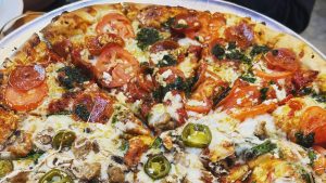 A vibrant dog park bathed in sunlight features abundant greenery with mature trees, offering ample shade for leisurely outdoor activities. In the distance is a well-equipped playground that presents an additional activity area. An inviting picnic table spreads out nearby, ideal for enjoying meals al fresco or taking relaxing breaks from play. A neatly-placed trash bin aids in maintaining the park's cleanliness for everyone's enjoyment. Dominating the scene is a generously wide dirt path gracefully curving through the expanse of grass, promising exploratory walks and lively fetch games.