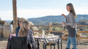 A vibrant dog park bathed in sunlight features abundant greenery with mature trees, offering ample shade for leisurely outdoor activities. In the distance is a well-equipped playground that presents an additional activity area. An inviting picnic table spreads out nearby, ideal for enjoying meals al fresco or taking relaxing breaks from play. A neatly-placed trash bin aids in maintaining the park's cleanliness for everyone's enjoyment. Dominating the scene is a generously wide dirt path gracefully curving through the expanse of grass, promising exploratory walks and lively fetch games.