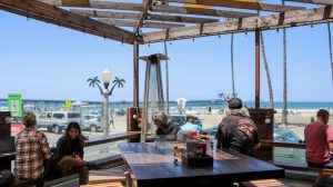 A vibrant dog park bathed in sunlight features abundant greenery with mature trees, offering ample shade for leisurely outdoor activities. In the distance is a well-equipped playground that presents an additional activity area. An inviting picnic table spreads out nearby, ideal for enjoying meals al fresco or taking relaxing breaks from play. A neatly-placed trash bin aids in maintaining the park's cleanliness for everyone's enjoyment. Dominating the scene is a generously wide dirt path gracefully curving through the expanse of grass, promising exploratory walks and lively fetch games.