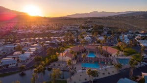 A vibrant dog park bathed in sunlight features abundant greenery with mature trees, offering ample shade for leisurely outdoor activities. In the distance is a well-equipped playground that presents an additional activity area. An inviting picnic table spreads out nearby, ideal for enjoying meals al fresco or taking relaxing breaks from play. A neatly-placed trash bin aids in maintaining the park's cleanliness for everyone's enjoyment. Dominating the scene is a generously wide dirt path gracefully curving through the expanse of grass, promising exploratory walks and lively fetch games.