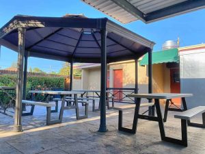 A vibrant dog park bathed in sunlight features abundant greenery with mature trees, offering ample shade for leisurely outdoor activities. In the distance is a well-equipped playground that presents an additional activity area. An inviting picnic table spreads out nearby, ideal for enjoying meals al fresco or taking relaxing breaks from play. A neatly-placed trash bin aids in maintaining the park's cleanliness for everyone's enjoyment. Dominating the scene is a generously wide dirt path gracefully curving through the expanse of grass, promising exploratory walks and lively fetch games.