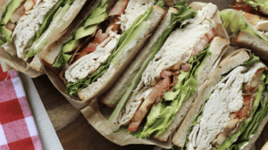 A vibrant dog park bathed in sunlight features abundant greenery with mature trees, offering ample shade for leisurely outdoor activities. In the distance is a well-equipped playground that presents an additional activity area. An inviting picnic table spreads out nearby, ideal for enjoying meals al fresco or taking relaxing breaks from play. A neatly-placed trash bin aids in maintaining the park's cleanliness for everyone's enjoyment. Dominating the scene is a generously wide dirt path gracefully curving through the expanse of grass, promising exploratory walks and lively fetch games.