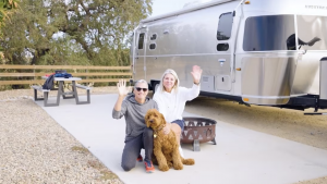 A vibrant dog park bathed in sunlight features abundant greenery with mature trees, offering ample shade for leisurely outdoor activities. In the distance is a well-equipped playground that presents an additional activity area. An inviting picnic table spreads out nearby, ideal for enjoying meals al fresco or taking relaxing breaks from play. A neatly-placed trash bin aids in maintaining the park's cleanliness for everyone's enjoyment. Dominating the scene is a generously wide dirt path gracefully curving through the expanse of grass, promising exploratory walks and lively fetch games.