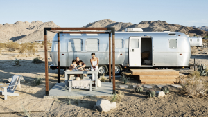 A vibrant dog park bathed in sunlight features abundant greenery with mature trees, offering ample shade for leisurely outdoor activities. In the distance is a well-equipped playground that presents an additional activity area. An inviting picnic table spreads out nearby, ideal for enjoying meals al fresco or taking relaxing breaks from play. A neatly-placed trash bin aids in maintaining the park's cleanliness for everyone's enjoyment. Dominating the scene is a generously wide dirt path gracefully curving through the expanse of grass, promising exploratory walks and lively fetch games.