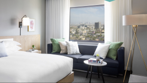 A vibrant dog park bathed in sunlight features abundant greenery with mature trees, offering ample shade for leisurely outdoor activities. In the distance is a well-equipped playground that presents an additional activity area. An inviting picnic table spreads out nearby, ideal for enjoying meals al fresco or taking relaxing breaks from play. A neatly-placed trash bin aids in maintaining the park's cleanliness for everyone's enjoyment. Dominating the scene is a generously wide dirt path gracefully curving through the expanse of grass, promising exploratory walks and lively fetch games.