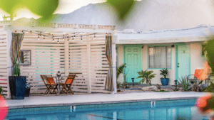 A vibrant dog park bathed in sunlight features abundant greenery with mature trees, offering ample shade for leisurely outdoor activities. In the distance is a well-equipped playground that presents an additional activity area. An inviting picnic table spreads out nearby, ideal for enjoying meals al fresco or taking relaxing breaks from play. A neatly-placed trash bin aids in maintaining the park's cleanliness for everyone's enjoyment. Dominating the scene is a generously wide dirt path gracefully curving through the expanse of grass, promising exploratory walks and lively fetch games.