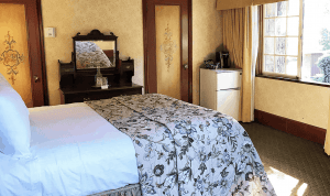 A vibrant dog park bathed in sunlight features abundant greenery with mature trees, offering ample shade for leisurely outdoor activities. In the distance is a well-equipped playground that presents an additional activity area. An inviting picnic table spreads out nearby, ideal for enjoying meals al fresco or taking relaxing breaks from play. A neatly-placed trash bin aids in maintaining the park's cleanliness for everyone's enjoyment. Dominating the scene is a generously wide dirt path gracefully curving through the expanse of grass, promising exploratory walks and lively fetch games.