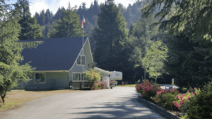 A vibrant dog park bathed in sunlight features abundant greenery with mature trees, offering ample shade for leisurely outdoor activities. In the distance is a well-equipped playground that presents an additional activity area. An inviting picnic table spreads out nearby, ideal for enjoying meals al fresco or taking relaxing breaks from play. A neatly-placed trash bin aids in maintaining the park's cleanliness for everyone's enjoyment. Dominating the scene is a generously wide dirt path gracefully curving through the expanse of grass, promising exploratory walks and lively fetch games.