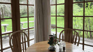 A vibrant dog park bathed in sunlight features abundant greenery with mature trees, offering ample shade for leisurely outdoor activities. In the distance is a well-equipped playground that presents an additional activity area. An inviting picnic table spreads out nearby, ideal for enjoying meals al fresco or taking relaxing breaks from play. A neatly-placed trash bin aids in maintaining the park's cleanliness for everyone's enjoyment. Dominating the scene is a generously wide dirt path gracefully curving through the expanse of grass, promising exploratory walks and lively fetch games.