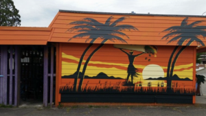 A vibrant dog park bathed in sunlight features abundant greenery with mature trees, offering ample shade for leisurely outdoor activities. In the distance is a well-equipped playground that presents an additional activity area. An inviting picnic table spreads out nearby, ideal for enjoying meals al fresco or taking relaxing breaks from play. A neatly-placed trash bin aids in maintaining the park's cleanliness for everyone's enjoyment. Dominating the scene is a generously wide dirt path gracefully curving through the expanse of grass, promising exploratory walks and lively fetch games.