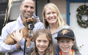 A vibrant dog park bathed in sunlight features abundant greenery with mature trees, offering ample shade for leisurely outdoor activities. In the distance is a well-equipped playground that presents an additional activity area. An inviting picnic table spreads out nearby, ideal for enjoying meals al fresco or taking relaxing breaks from play. A neatly-placed trash bin aids in maintaining the park's cleanliness for everyone's enjoyment. Dominating the scene is a generously wide dirt path gracefully curving through the expanse of grass, promising exploratory walks and lively fetch games.
