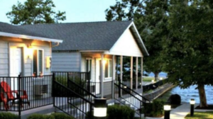 A vibrant dog park bathed in sunlight features abundant greenery with mature trees, offering ample shade for leisurely outdoor activities. In the distance is a well-equipped playground that presents an additional activity area. An inviting picnic table spreads out nearby, ideal for enjoying meals al fresco or taking relaxing breaks from play. A neatly-placed trash bin aids in maintaining the park's cleanliness for everyone's enjoyment. Dominating the scene is a generously wide dirt path gracefully curving through the expanse of grass, promising exploratory walks and lively fetch games.