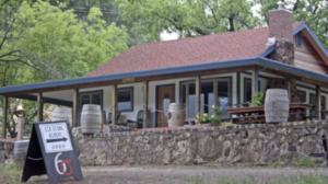 A vibrant dog park bathed in sunlight features abundant greenery with mature trees, offering ample shade for leisurely outdoor activities. In the distance is a well-equipped playground that presents an additional activity area. An inviting picnic table spreads out nearby, ideal for enjoying meals al fresco or taking relaxing breaks from play. A neatly-placed trash bin aids in maintaining the park's cleanliness for everyone's enjoyment. Dominating the scene is a generously wide dirt path gracefully curving through the expanse of grass, promising exploratory walks and lively fetch games.