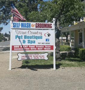 A vibrant dog park bathed in sunlight features abundant greenery with mature trees, offering ample shade for leisurely outdoor activities. In the distance is a well-equipped playground that presents an additional activity area. An inviting picnic table spreads out nearby, ideal for enjoying meals al fresco or taking relaxing breaks from play. A neatly-placed trash bin aids in maintaining the park's cleanliness for everyone's enjoyment. Dominating the scene is a generously wide dirt path gracefully curving through the expanse of grass, promising exploratory walks and lively fetch games.