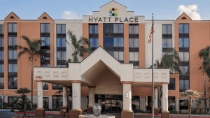 A vibrant dog park bathed in sunlight features abundant greenery with mature trees, offering ample shade for leisurely outdoor activities. In the distance is a well-equipped playground that presents an additional activity area. An inviting picnic table spreads out nearby, ideal for enjoying meals al fresco or taking relaxing breaks from play. A neatly-placed trash bin aids in maintaining the park's cleanliness for everyone's enjoyment. Dominating the scene is a generously wide dirt path gracefully curving through the expanse of grass, promising exploratory walks and lively fetch games.