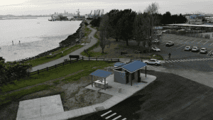 A vibrant dog park bathed in sunlight features abundant greenery with mature trees, offering ample shade for leisurely outdoor activities. In the distance is a well-equipped playground that presents an additional activity area. An inviting picnic table spreads out nearby, ideal for enjoying meals al fresco or taking relaxing breaks from play. A neatly-placed trash bin aids in maintaining the park's cleanliness for everyone's enjoyment. Dominating the scene is a generously wide dirt path gracefully curving through the expanse of grass, promising exploratory walks and lively fetch games.