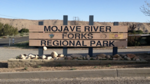 A vibrant dog park bathed in sunlight features abundant greenery with mature trees, offering ample shade for leisurely outdoor activities. In the distance is a well-equipped playground that presents an additional activity area. An inviting picnic table spreads out nearby, ideal for enjoying meals al fresco or taking relaxing breaks from play. A neatly-placed trash bin aids in maintaining the park's cleanliness for everyone's enjoyment. Dominating the scene is a generously wide dirt path gracefully curving through the expanse of grass, promising exploratory walks and lively fetch games.