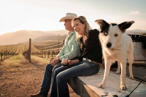 A vibrant dog park bathed in sunlight features abundant greenery with mature trees, offering ample shade for leisurely outdoor activities. In the distance is a well-equipped playground that presents an additional activity area. An inviting picnic table spreads out nearby, ideal for enjoying meals al fresco or taking relaxing breaks from play. A neatly-placed trash bin aids in maintaining the park's cleanliness for everyone's enjoyment. Dominating the scene is a generously wide dirt path gracefully curving through the expanse of grass, promising exploratory walks and lively fetch games.