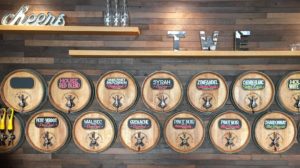 A vibrant dog park bathed in sunlight features abundant greenery with mature trees, offering ample shade for leisurely outdoor activities. In the distance is a well-equipped playground that presents an additional activity area. An inviting picnic table spreads out nearby, ideal for enjoying meals al fresco or taking relaxing breaks from play. A neatly-placed trash bin aids in maintaining the park's cleanliness for everyone's enjoyment. Dominating the scene is a generously wide dirt path gracefully curving through the expanse of grass, promising exploratory walks and lively fetch games.