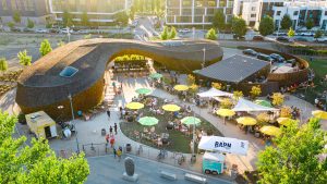 A vibrant dog park bathed in sunlight features abundant greenery with mature trees, offering ample shade for leisurely outdoor activities. In the distance is a well-equipped playground that presents an additional activity area. An inviting picnic table spreads out nearby, ideal for enjoying meals al fresco or taking relaxing breaks from play. A neatly-placed trash bin aids in maintaining the park's cleanliness for everyone's enjoyment. Dominating the scene is a generously wide dirt path gracefully curving through the expanse of grass, promising exploratory walks and lively fetch games.