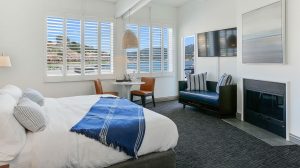 A vibrant dog park bathed in sunlight features abundant greenery with mature trees, offering ample shade for leisurely outdoor activities. In the distance is a well-equipped playground that presents an additional activity area. An inviting picnic table spreads out nearby, ideal for enjoying meals al fresco or taking relaxing breaks from play. A neatly-placed trash bin aids in maintaining the park's cleanliness for everyone's enjoyment. Dominating the scene is a generously wide dirt path gracefully curving through the expanse of grass, promising exploratory walks and lively fetch games.