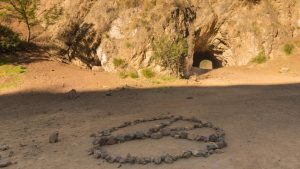 A vibrant dog park bathed in sunlight features abundant greenery with mature trees, offering ample shade for leisurely outdoor activities. In the distance is a well-equipped playground that presents an additional activity area. An inviting picnic table spreads out nearby, ideal for enjoying meals al fresco or taking relaxing breaks from play. A neatly-placed trash bin aids in maintaining the park's cleanliness for everyone's enjoyment. Dominating the scene is a generously wide dirt path gracefully curving through the expanse of grass, promising exploratory walks and lively fetch games.