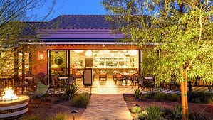 A vibrant dog park bathed in sunlight features abundant greenery with mature trees, offering ample shade for leisurely outdoor activities. In the distance is a well-equipped playground that presents an additional activity area. An inviting picnic table spreads out nearby, ideal for enjoying meals al fresco or taking relaxing breaks from play. A neatly-placed trash bin aids in maintaining the park's cleanliness for everyone's enjoyment. Dominating the scene is a generously wide dirt path gracefully curving through the expanse of grass, promising exploratory walks and lively fetch games.