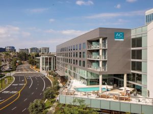 AC Hotel Irvine with balconies and an outdoor pool sits by a busy road with palm trees; nearby parks offer dog-friendly spaces.