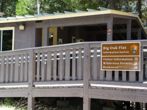 A vibrant dog park bathed in sunlight features abundant greenery with mature trees, offering ample shade for leisurely outdoor activities. In the distance is a well-equipped playground that presents an additional activity area. An inviting picnic table spreads out nearby, ideal for enjoying meals al fresco or taking relaxing breaks from play. A neatly-placed trash bin aids in maintaining the park's cleanliness for everyone's enjoyment. Dominating the scene is a generously wide dirt path gracefully curving through the expanse of grass, promising exploratory walks and lively fetch games.