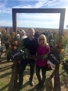A vibrant dog park bathed in sunlight features abundant greenery with mature trees, offering ample shade for leisurely outdoor activities. In the distance is a well-equipped playground that presents an additional activity area. An inviting picnic table spreads out nearby, ideal for enjoying meals al fresco or taking relaxing breaks from play. A neatly-placed trash bin aids in maintaining the park's cleanliness for everyone's enjoyment. Dominating the scene is a generously wide dirt path gracefully curving through the expanse of grass, promising exploratory walks and lively fetch games.
