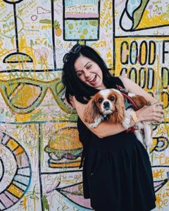 A vibrant dog park bathed in sunlight features abundant greenery with mature trees, offering ample shade for leisurely outdoor activities. In the distance is a well-equipped playground that presents an additional activity area. An inviting picnic table spreads out nearby, ideal for enjoying meals al fresco or taking relaxing breaks from play. A neatly-placed trash bin aids in maintaining the park's cleanliness for everyone's enjoyment. Dominating the scene is a generously wide dirt path gracefully curving through the expanse of grass, promising exploratory walks and lively fetch games.