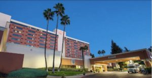 A vibrant dog park bathed in sunlight features abundant greenery with mature trees, offering ample shade for leisurely outdoor activities. In the distance is a well-equipped playground that presents an additional activity area. An inviting picnic table spreads out nearby, ideal for enjoying meals al fresco or taking relaxing breaks from play. A neatly-placed trash bin aids in maintaining the park's cleanliness for everyone's enjoyment. Dominating the scene is a generously wide dirt path gracefully curving through the expanse of grass, promising exploratory walks and lively fetch games.