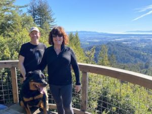 A vibrant dog park bathed in sunlight features abundant greenery with mature trees, offering ample shade for leisurely outdoor activities. In the distance is a well-equipped playground that presents an additional activity area. An inviting picnic table spreads out nearby, ideal for enjoying meals al fresco or taking relaxing breaks from play. A neatly-placed trash bin aids in maintaining the park's cleanliness for everyone's enjoyment. Dominating the scene is a generously wide dirt path gracefully curving through the expanse of grass, promising exploratory walks and lively fetch games.