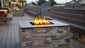 A vibrant dog park bathed in sunlight features abundant greenery with mature trees, offering ample shade for leisurely outdoor activities. In the distance is a well-equipped playground that presents an additional activity area. An inviting picnic table spreads out nearby, ideal for enjoying meals al fresco or taking relaxing breaks from play. A neatly-placed trash bin aids in maintaining the park's cleanliness for everyone's enjoyment. Dominating the scene is a generously wide dirt path gracefully curving through the expanse of grass, promising exploratory walks and lively fetch games.