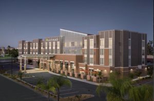 A vibrant dog park bathed in sunlight features abundant greenery with mature trees, offering ample shade for leisurely outdoor activities. In the distance is a well-equipped playground that presents an additional activity area. An inviting picnic table spreads out nearby, ideal for enjoying meals al fresco or taking relaxing breaks from play. A neatly-placed trash bin aids in maintaining the park's cleanliness for everyone's enjoyment. Dominating the scene is a generously wide dirt path gracefully curving through the expanse of grass, promising exploratory walks and lively fetch games.