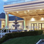 A vibrant dog park bathed in sunlight features abundant greenery with mature trees, offering ample shade for leisurely outdoor activities. In the distance is a well-equipped playground that presents an additional activity area. An inviting picnic table spreads out nearby, ideal for enjoying meals al fresco or taking relaxing breaks from play. A neatly-placed trash bin aids in maintaining the park's cleanliness for everyone's enjoyment. Dominating the scene is a generously wide dirt path gracefully curving through the expanse of grass, promising exploratory walks and lively fetch games.