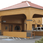 A vibrant dog park bathed in sunlight features abundant greenery with mature trees, offering ample shade for leisurely outdoor activities. In the distance is a well-equipped playground that presents an additional activity area. An inviting picnic table spreads out nearby, ideal for enjoying meals al fresco or taking relaxing breaks from play. A neatly-placed trash bin aids in maintaining the park's cleanliness for everyone's enjoyment. Dominating the scene is a generously wide dirt path gracefully curving through the expanse of grass, promising exploratory walks and lively fetch games.