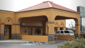 A vibrant dog park bathed in sunlight features abundant greenery with mature trees, offering ample shade for leisurely outdoor activities. In the distance is a well-equipped playground that presents an additional activity area. An inviting picnic table spreads out nearby, ideal for enjoying meals al fresco or taking relaxing breaks from play. A neatly-placed trash bin aids in maintaining the park's cleanliness for everyone's enjoyment. Dominating the scene is a generously wide dirt path gracefully curving through the expanse of grass, promising exploratory walks and lively fetch games.