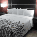 A vibrant dog park bathed in sunlight features abundant greenery with mature trees, offering ample shade for leisurely outdoor activities. In the distance is a well-equipped playground that presents an additional activity area. An inviting picnic table spreads out nearby, ideal for enjoying meals al fresco or taking relaxing breaks from play. A neatly-placed trash bin aids in maintaining the park's cleanliness for everyone's enjoyment. Dominating the scene is a generously wide dirt path gracefully curving through the expanse of grass, promising exploratory walks and lively fetch games.