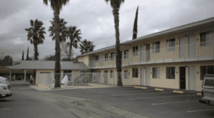 A vibrant dog park bathed in sunlight features abundant greenery with mature trees, offering ample shade for leisurely outdoor activities. In the distance is a well-equipped playground that presents an additional activity area. An inviting picnic table spreads out nearby, ideal for enjoying meals al fresco or taking relaxing breaks from play. A neatly-placed trash bin aids in maintaining the park's cleanliness for everyone's enjoyment. Dominating the scene is a generously wide dirt path gracefully curving through the expanse of grass, promising exploratory walks and lively fetch games.