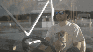 A vibrant dog park bathed in sunlight features abundant greenery with mature trees, offering ample shade for leisurely outdoor activities. In the distance is a well-equipped playground that presents an additional activity area. An inviting picnic table spreads out nearby, ideal for enjoying meals al fresco or taking relaxing breaks from play. A neatly-placed trash bin aids in maintaining the park's cleanliness for everyone's enjoyment. Dominating the scene is a generously wide dirt path gracefully curving through the expanse of grass, promising exploratory walks and lively fetch games.